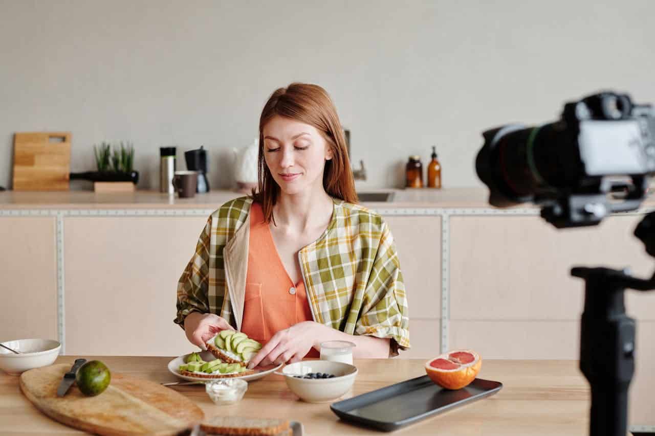 hero-img-01 Caucasian woman preparing breakfast with avocado toast while recording a cooking vlog in a kitchen.